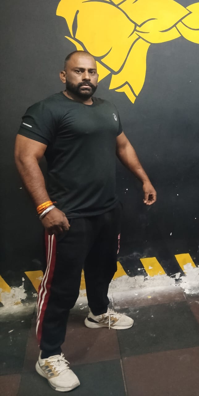 PAHLAWAN JI in black t-shirt standing in front of yellow lion wall, strength coach portrait